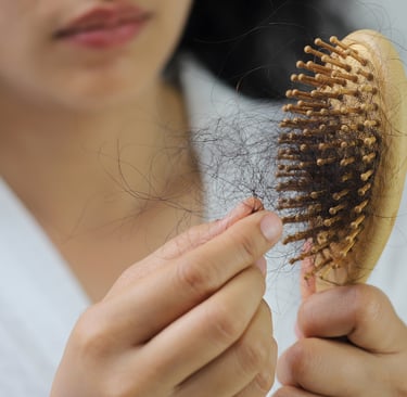 Woman in a white robe holding a wooden hairbrush with tangled hair loss strands after brushing.