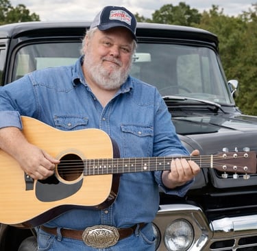 Country singer Bubba Bohacks plays an acoustic guitar in front of a classic black Ford truck.