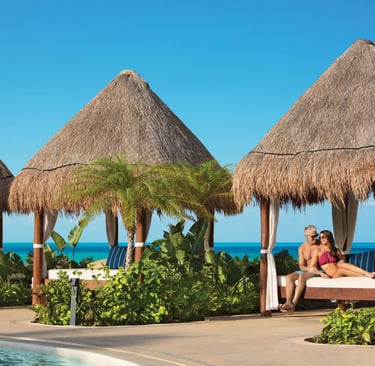 A couple relaxes in a luxury beachfront cabana with thatched roofs at a tropical resort.