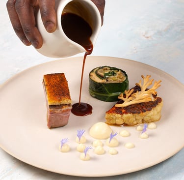 Chef pouring dark jus over a gourmet plate of roasted pork belly, stuffed cabbage, and cauliflower steak.
