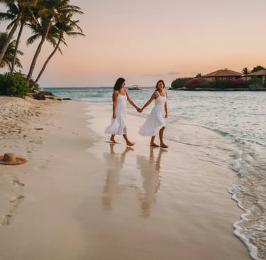Two women in white dresses holding hands while walking along a tropical beach at sunset.