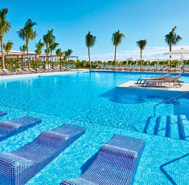 Luxury tropical resort swimming pool with submerged lounge chairs, palm trees, and sunny blue sky.