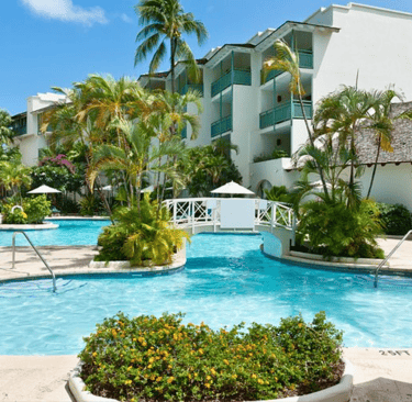 Luxury tropical resort pool with a white footbridge, palm trees, and bright blue water under a clear sky.