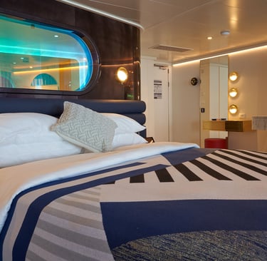 Modern luxury cruise ship cabin featuring a plush bed with geometric bedding and an oval porthole window.