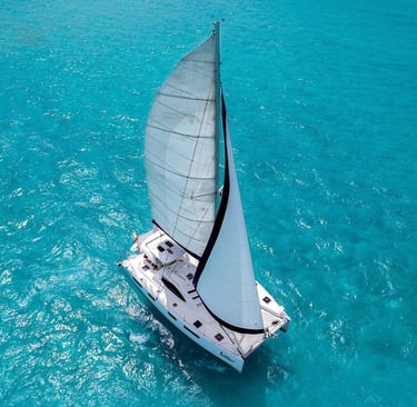 An aerial view of a white sailing catamaran cruising through clear turquoise tropical ocean water.