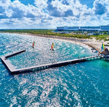 Aerial view of a luxury beachfront resort with a long wooden pier, turquoise water, and windsurfers.