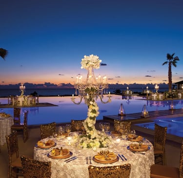 Luxury outdoor wedding reception by a pool at sunset with gold chairs and floral candelabra centerpieces.