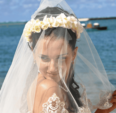 A bride with a flower crown and white lace veil during a tropical beach wedding ceremony.