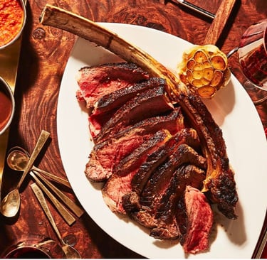 Sliced medium-rare tomahawk ribeye steak served with roasted garlic, sauces, and red wine.