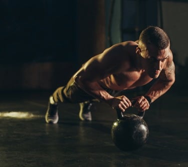 Shirtless muscular man performing a kettlebell pushup for strength training in a dark gym.