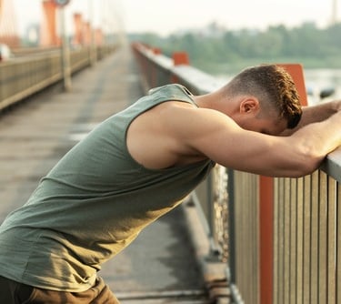Athletic man in tank top leaning on a bridge railing while resting after an outdoor workout.