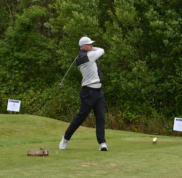 A male golfer in a white cap follows through on a drive during a tournament at a lush green golf course.