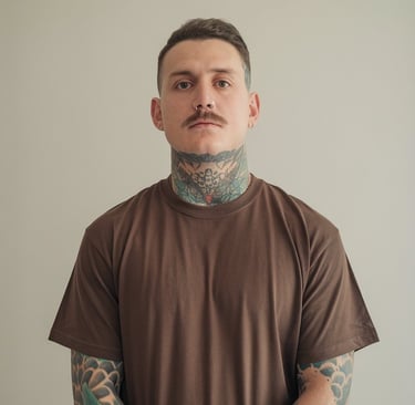 Portrait d’un homme tatoué, debout devant un fond neutre. Il a une moustache, un piercing au nez,