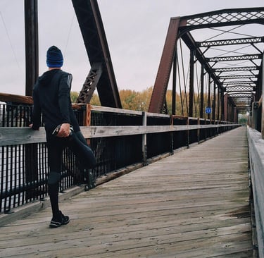a man in a blue hat and a blue jacket is standing on a bridge. he is warming up for a run to maintai