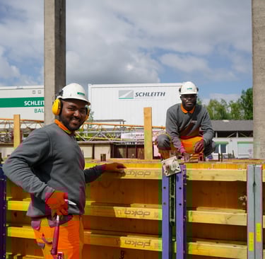 Zwei Bauarbeiter in Schutzkleidung und Helmen arbeiten lächelnd auf einer Baustelle.