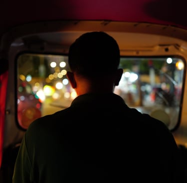 Silhouette of a tuk‑tuk driver in Phnom Penh facing blurred city lights at night, By ACAT Photos.