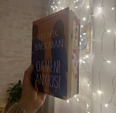Mână ținând cartea 'Oameni anxioși' de Fredrik Backman pe un fundal de luminițe de basm.