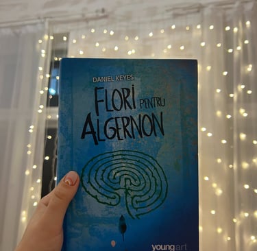 Mână ținând cartea 'Flori pentru Algernon' de Daniel Keyes pe un fundal de luminițe de basm.