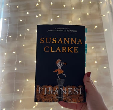 Mână ținând cartea 'Piranesi' de Susanna Clarke pe un fundal de luminițe de basm.
