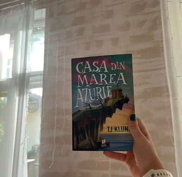 Mână ținând cartea 'Casa din Marea Azurie' de TJ Klune pe un fundal de luminițe de basm.
