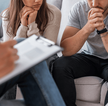 2 people, partially visible, sitting on a couch across from someone taking notes on a clipboard