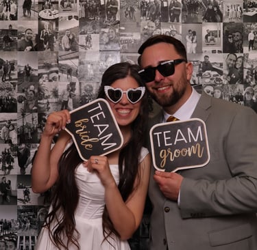 Bride and groom posing in a photo booth with fun signs and heart-shaped sunglasses.