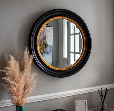 Wall with a mirror featuring a black frame and gold lining, above a shelf with decorative pieces