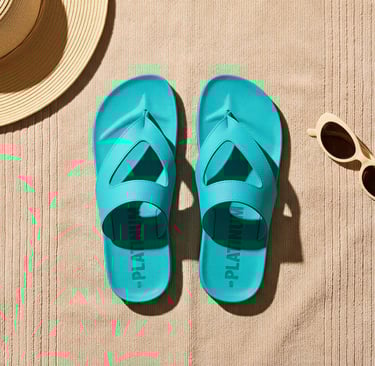 Turquoise flip flops on a beach towel with a straw hat and sunglasses on sand.