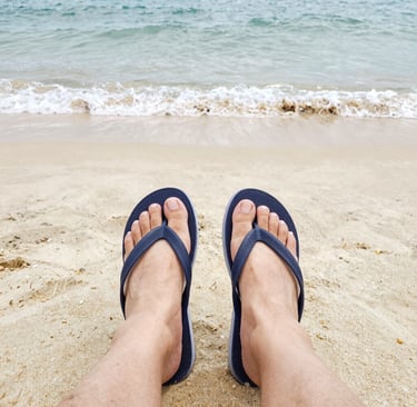 Person wearing navy blue flip flops on a sandy tropical beach with ocean waves.