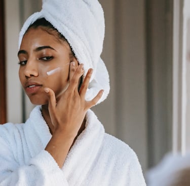 a woman in a white robe is looking at her face with a towel on her head applying face cream