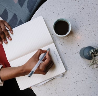 A hand journaling on a notebook that is placed on a table with a cup of tea.