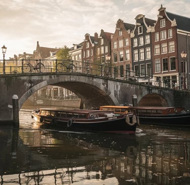 Canal tour boats cruising under a stone bridge in Amsterdam at sunset with historic gabled houses.