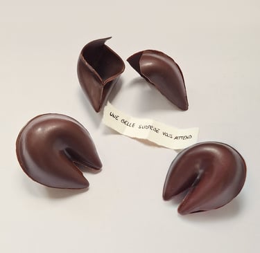 Handmade dark chocolate fortune cookies on a white surface with a paper message in French.