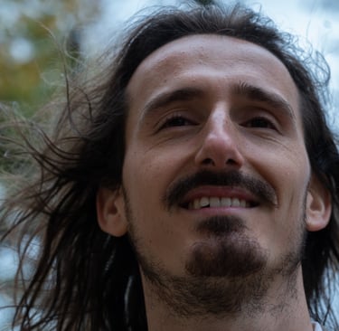 Smiling man with long brown hair and a goatee posing outdoors with wind blowing through his hair.
