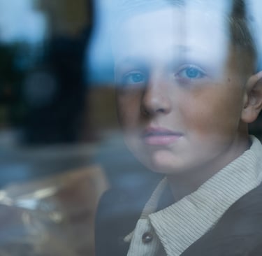 A young boy looking through a glass window with a pensive expression.