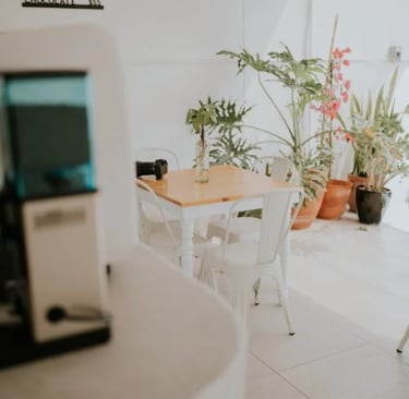 Varanda clara com mesas de madeira e cadeiras brancas, decorada com plantas em vasos grandes e luz n