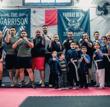 Adults and children group posing in a Combat Budo martial arts dojo for self-defense training.