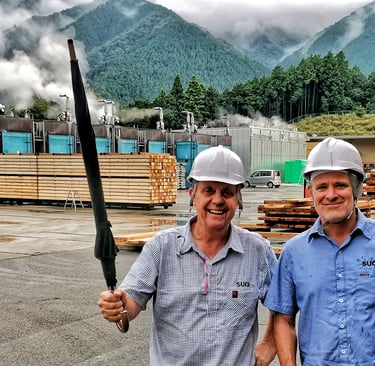 Two men in hard hats stand before a Japanese sawmill with wood stacks and misty mountains.