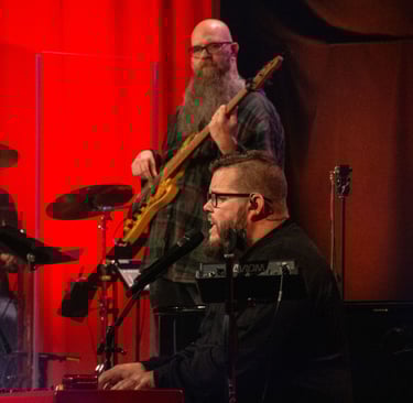 A worship team playing music on a church stage, with drums, bass guitar, piano, and vocalists
