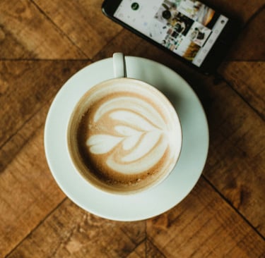Taza de café con latte art sobre mesa de madera junto a un teléfono móvil, reflejando el ritual y es