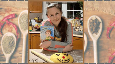 A woman smiling, holding a children's book titled "Living Dough," with a dough face on the table.