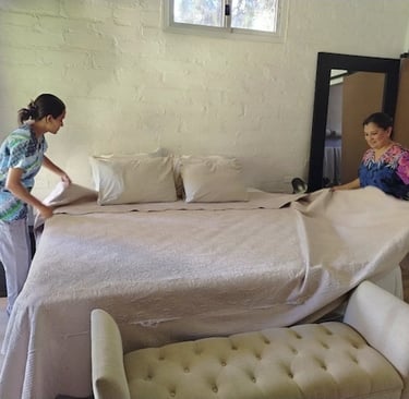 Two professional housekeepers in uniforms making a large bed with fresh linens in a modern bedroom.