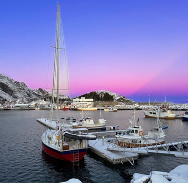 Småbåthavn i Moskenes i Lofoten med seilbåt, fiskebåter og vinterfjell i bakgrunnen i rosa og blått 