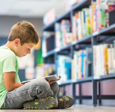 imagem mostra uma criança de 10 anos aproximadamente lendo um livro sentado no chão da biblioteca