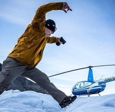 A photographer in hiking gear and crampons leaps across an icy glacier near a blue rescue helicopter.