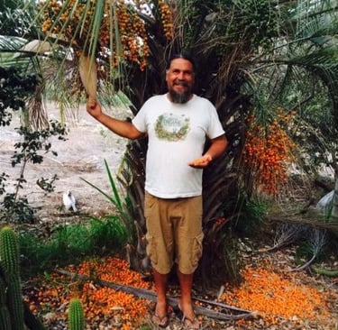 Man in white shirt and tan shorts standing in front of a palm with many orange fruit