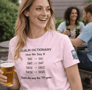 A smiling woman in a pink Dublin Dictionary t-shirt holding a beer at an outdoor patio.