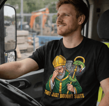 A smiling man on a couch, holding a white mug and remote, wearing a t-shirt featuring a cartoon St. Patrick looking