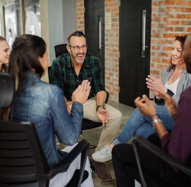 A diverse group of adults sit in a circle, smiling and talking during a relaxed group discussion or