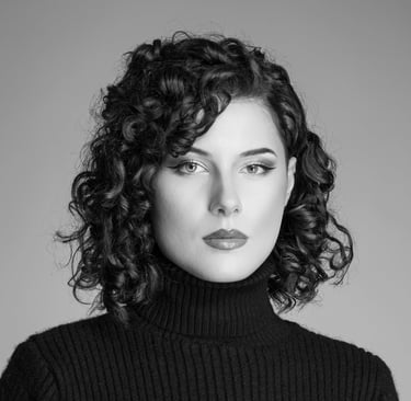  Ein Schwarz-Weiß-Porträt einer Frau mit kurzen, lockigen Haaren. Sie trägt einen dunklen Rollkragenpullover und hat einen au